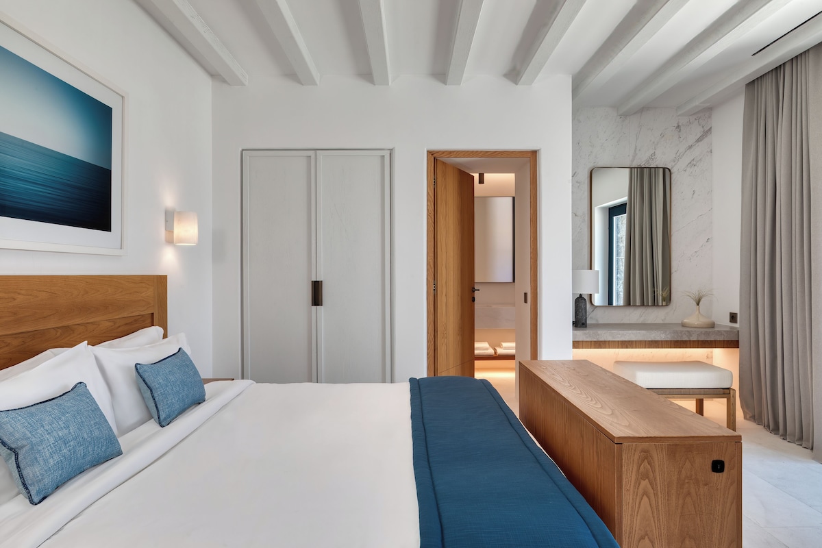 A modern hotel room with a large bed, a wooden bench, a bedside table, and a bathroom visible through an open door.