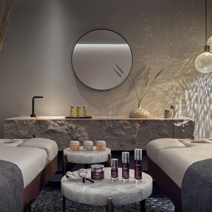 A modern spa room with two beds, a marble counter with skincare products, a round mirror, and a wooden shelf with towels.