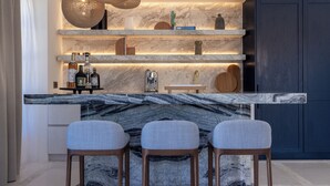 A modern kitchen with a marble countertop, three bar stools, and a built-in shelf with various items.