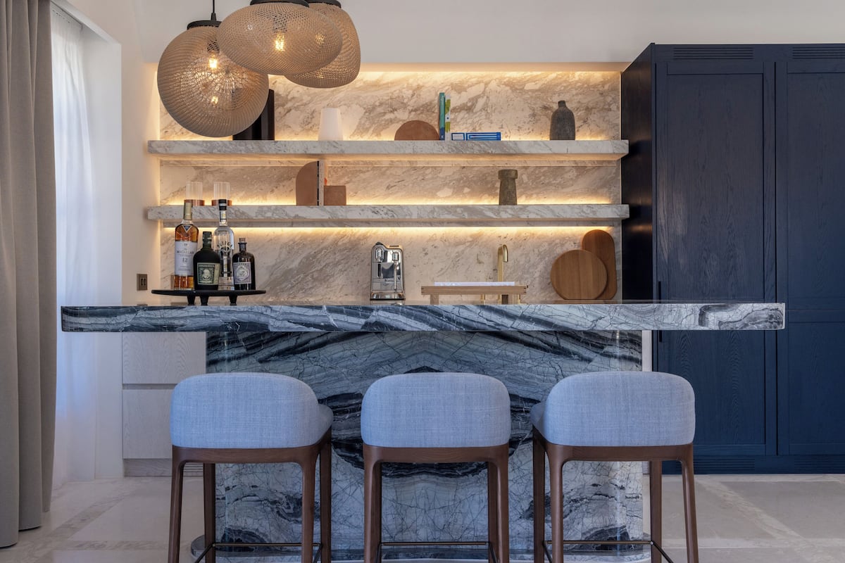 A modern kitchen with a marble countertop, three bar stools, and a built-in shelf with various items.