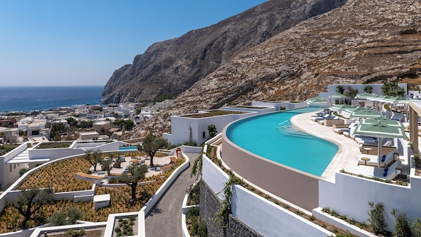 A resort with a curved pool, white buildings, and a view of the sea and mountains.