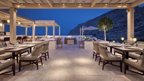 An outdoor dining area with tables and chairs, a pergola structure, and a view of the sea and mountains at dusk.