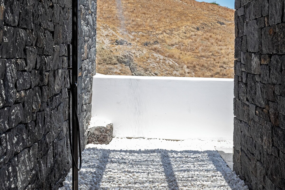A stone-walled outdoor shower area with a water spout, surrounded by white pebbles and a clear view of a hillside.