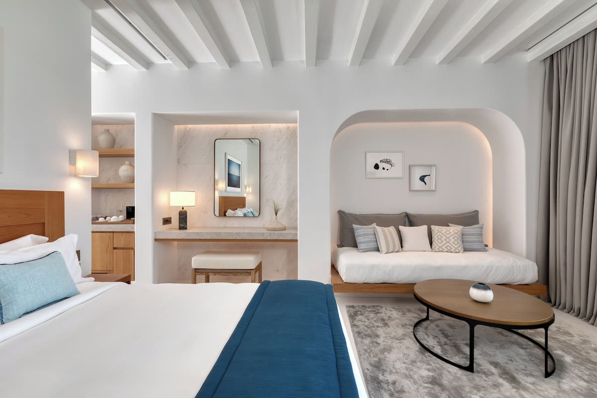 A modern hotel room with a large bed, a sofa, a small table, and a bathroom area with a mirror and vanity.
