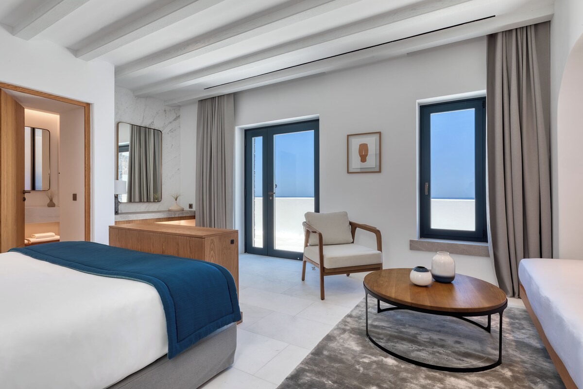 Et moderne hotelværelse med en stor seng, et siddeområde, et lille bord, et badeværelse med et spejl og store vinduer med udsigt.