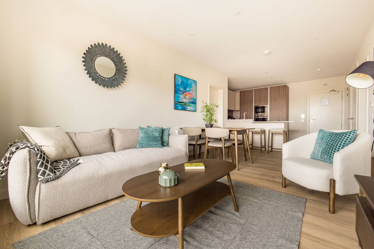 A modern living room with a sofa, coffee table, and armchairs. A dining area with a table and chairs is visible in the background.
