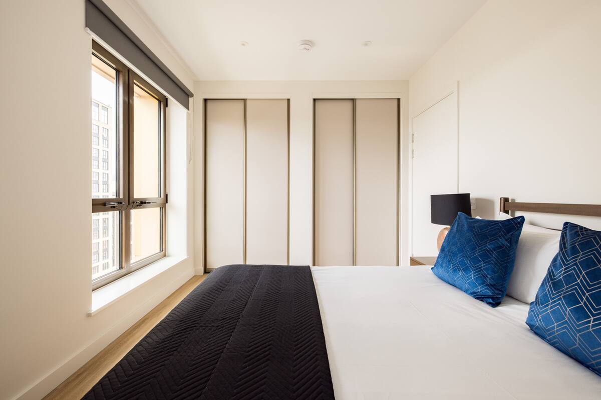 A hotel room with a large bed, a window with blinds, a bedside table with a lamp, and a view of the city through the window.