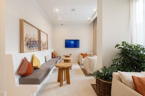 A modern lounge area with a sofa, wooden stools, a television, and a large plant.
