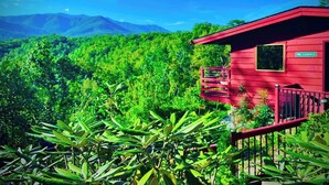 A red cabin with a balcony, surrounded by lush greenery and mountains in the background. - Stunning Mtn View, True Log Cabin, Romance Getaway, Rare Find (Gatlinburg)