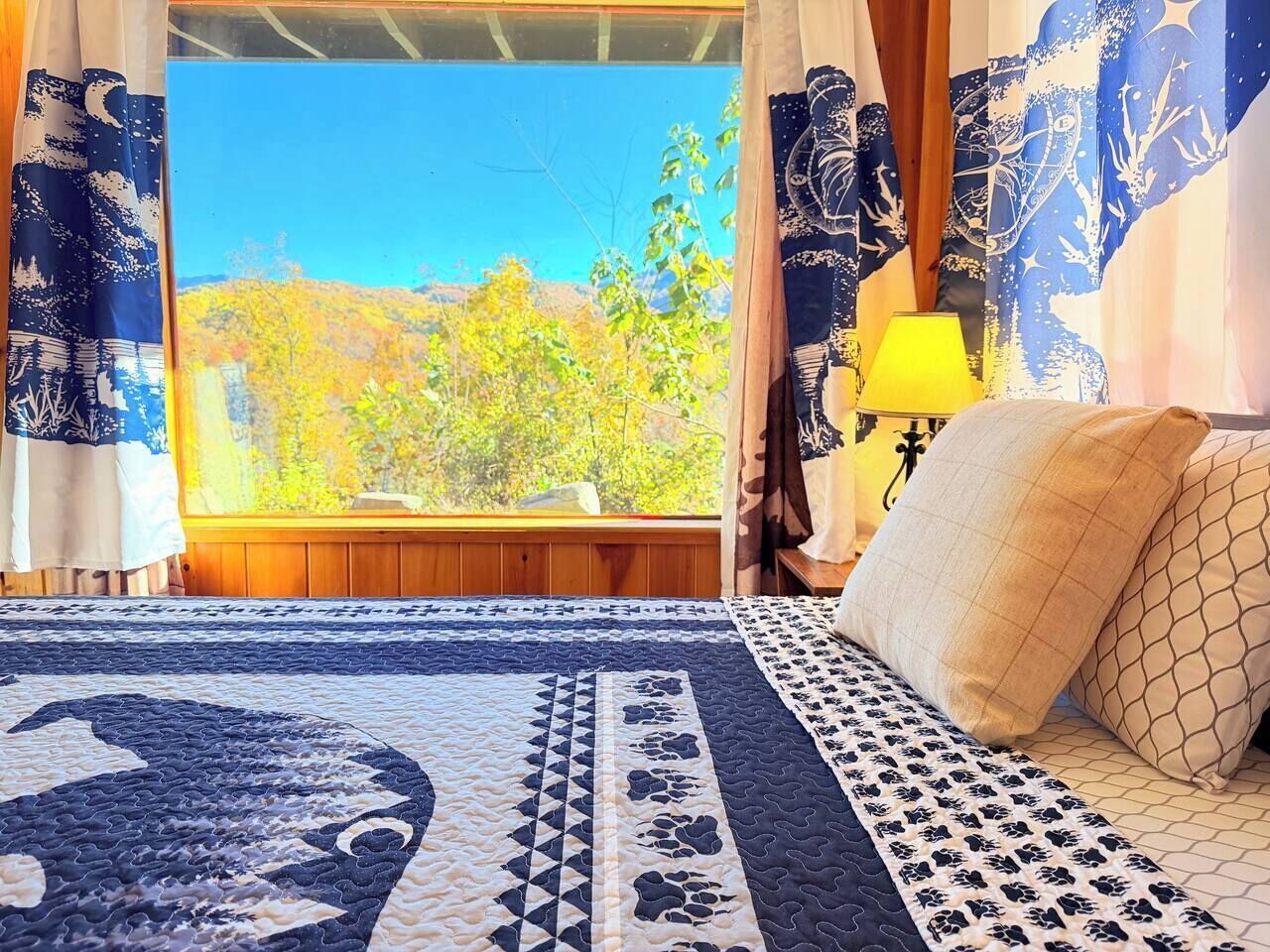 Un dormitorio con un ventanal grande, una cama con ropa de cama estampada, una mesita de noche con una lámpara y cortinas con diseño celestial.