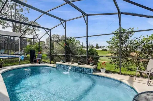 A swimming pool with a waterfall feature, surrounded by a covered area with chairs and potted plants. - Golf Course Retreat (Haines City)
