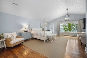 Quarto com cama de dossel, um espaço de estar com uma poltrona e uma mesinha, janela com cortinas e ventoinha no teto.