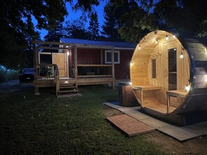 A wooden cabin with a large circular room, a small porch with benches, and a car parked nearby. - Cardinal Landing – Private Luxury Getaway with Sauna, Cold Plunge & Nature (Southampton)