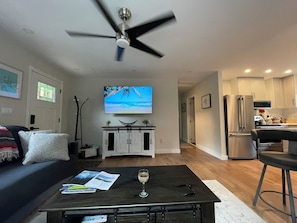 Una sala moderna con televisor de pantalla plana, una mesa de centro con revistas y una copa de vino, un ventilador de techo y una zona de cocina con electrodomésticos de acero inoxidable.