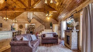 A rustic log cabin interior with a wooden staircase, a fireplace, and a cozy seating area with two sofas.