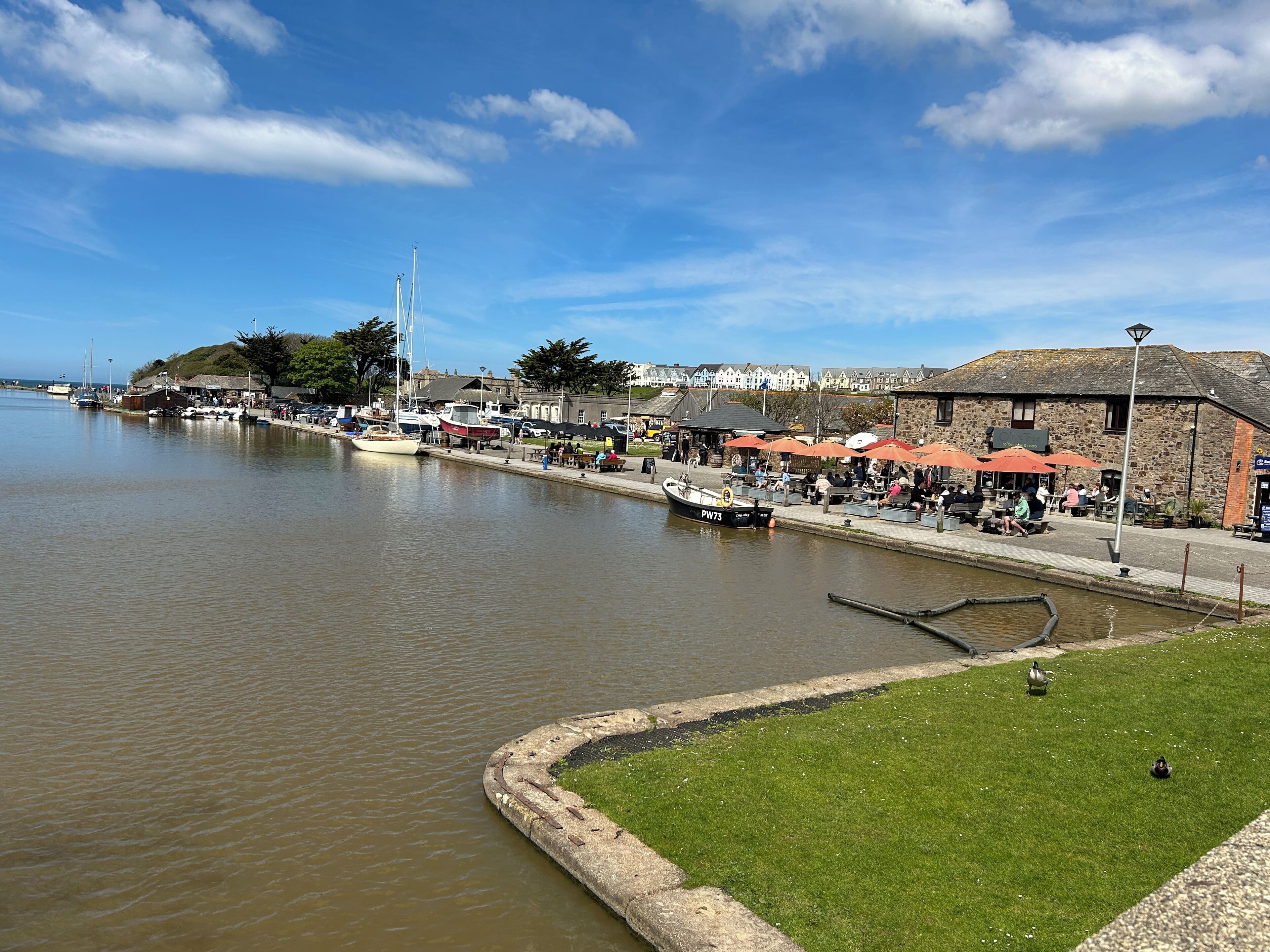 A riverside town with a stone building, a grassy area, and a dock with boats.