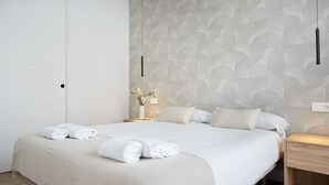 A neatly made bed with white linens and rolled towels, a bedside table with a vase of flowers, and a wall with a circular pattern.