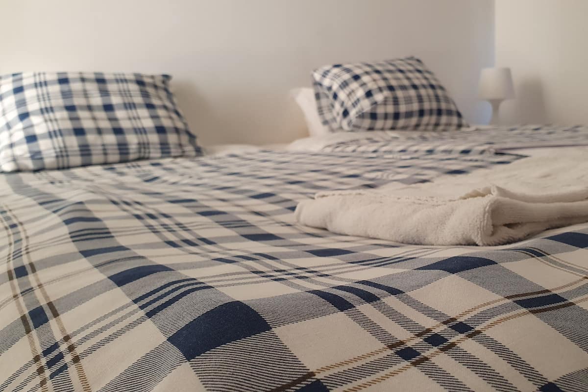 Una cama con una colcha a cuadros y dos almohadas, una toalla blanca doblada y una mesita de noche con una lámpara.