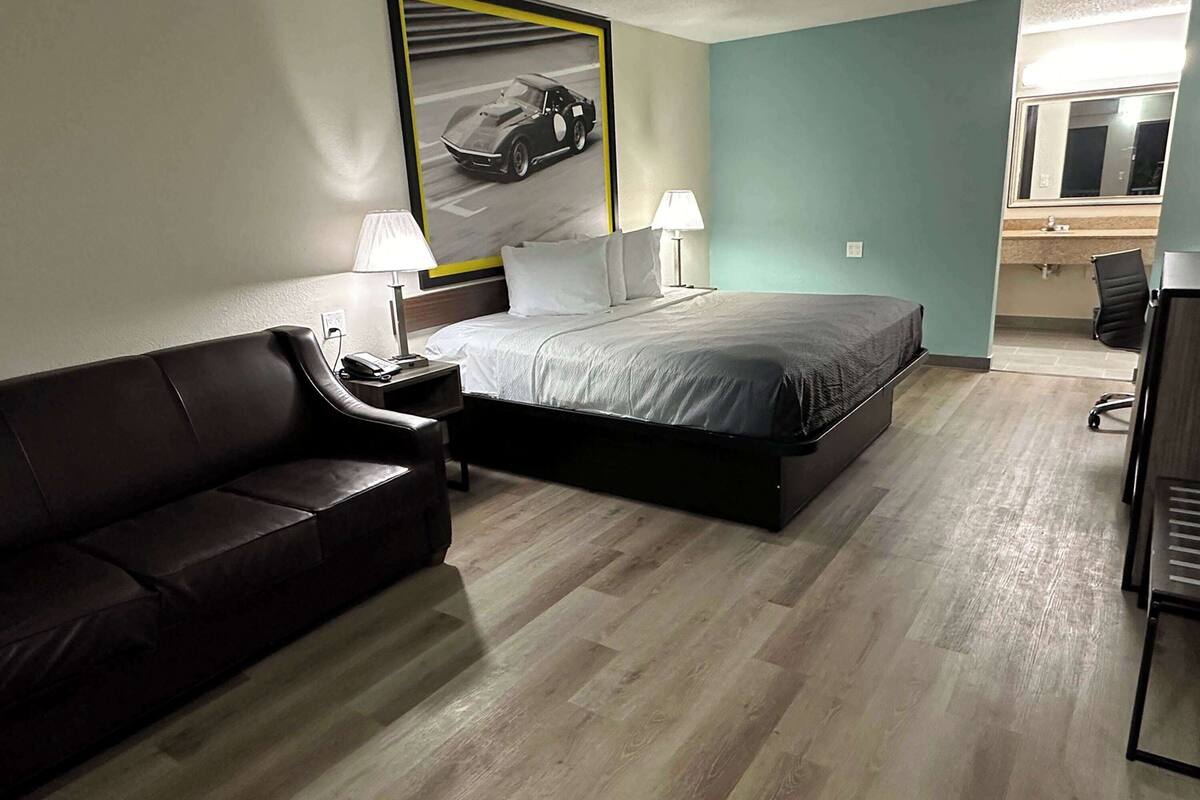 Ett hotellrum med en säng, en mörk lädersoffa, ett skrivbord med en lampa, en väggmonterad TV och ett badrum som syns genom en öppen dörr.