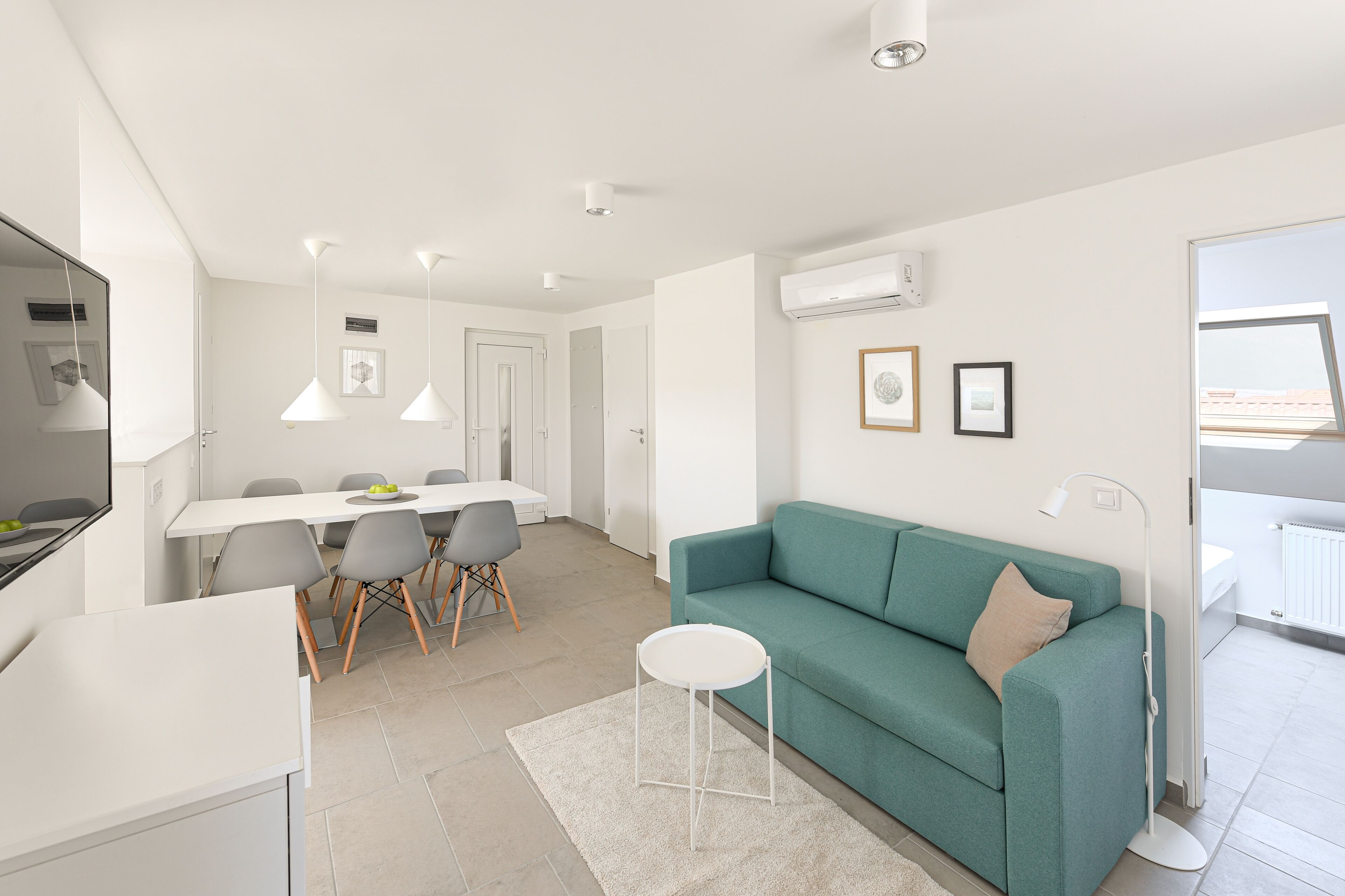 Una sala de estar moderna con un sofá color turquesa, una mesita redonda pequeña y un comedor con una mesa blanca y sillas grises.