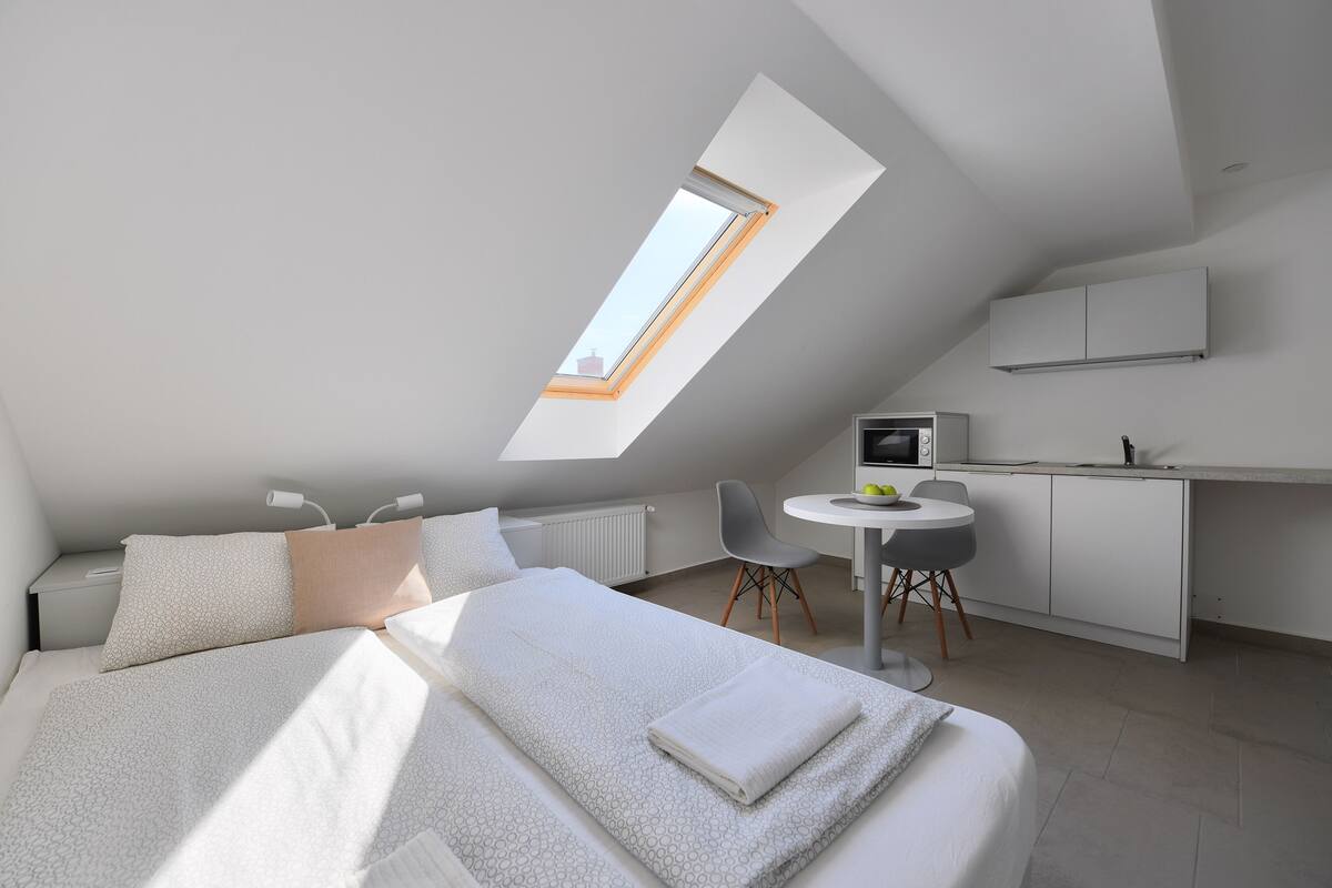 Quarto moderno com claraboia, cama com roupa de cama branca, pequena escrivaninha com cadeira e área de cozinha compacta.