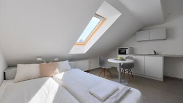 Ein modernes Schlafzimmer mit Dachfenster, einem Bett mit weiĂer BettwĂ€sche, einem kleinen Schreibtisch mit Stuhl und einer KĂŒchenzeile.
