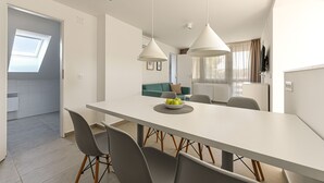 A modern dining area with a white table, grey chairs, and pendant lighting. A bowl of fruit is on the table. - Villa Oliver Hévíz 1 (Hévíz)