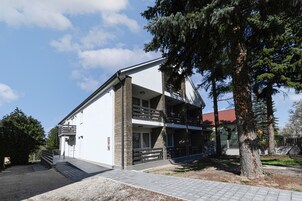 Um edifício de dois andares com varanda no segundo andar, telhado vermelho e entrada com calçamento de brita.