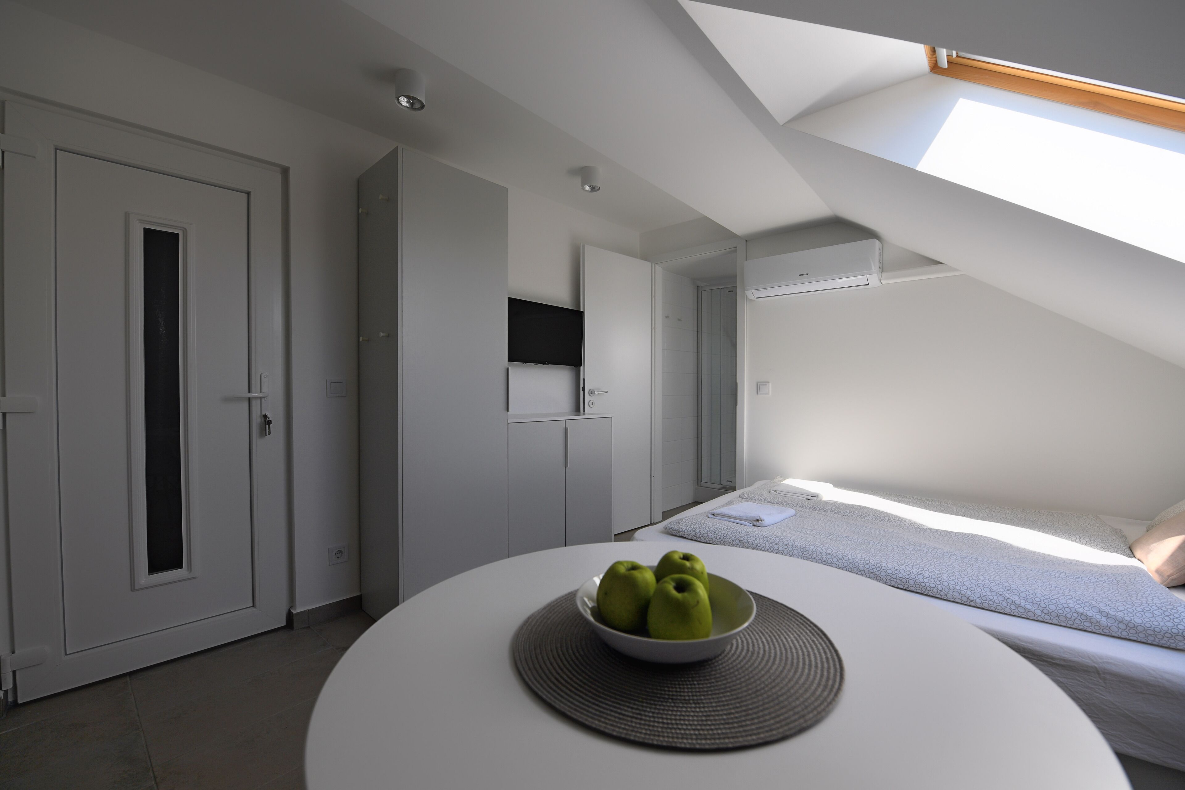 Une chambre à coucher moderne avec une lucarne, une table ronde sur laquelle se trouve un saladier rempli de pommes, et un lit recouvert d’une couvre-lit à motifs.