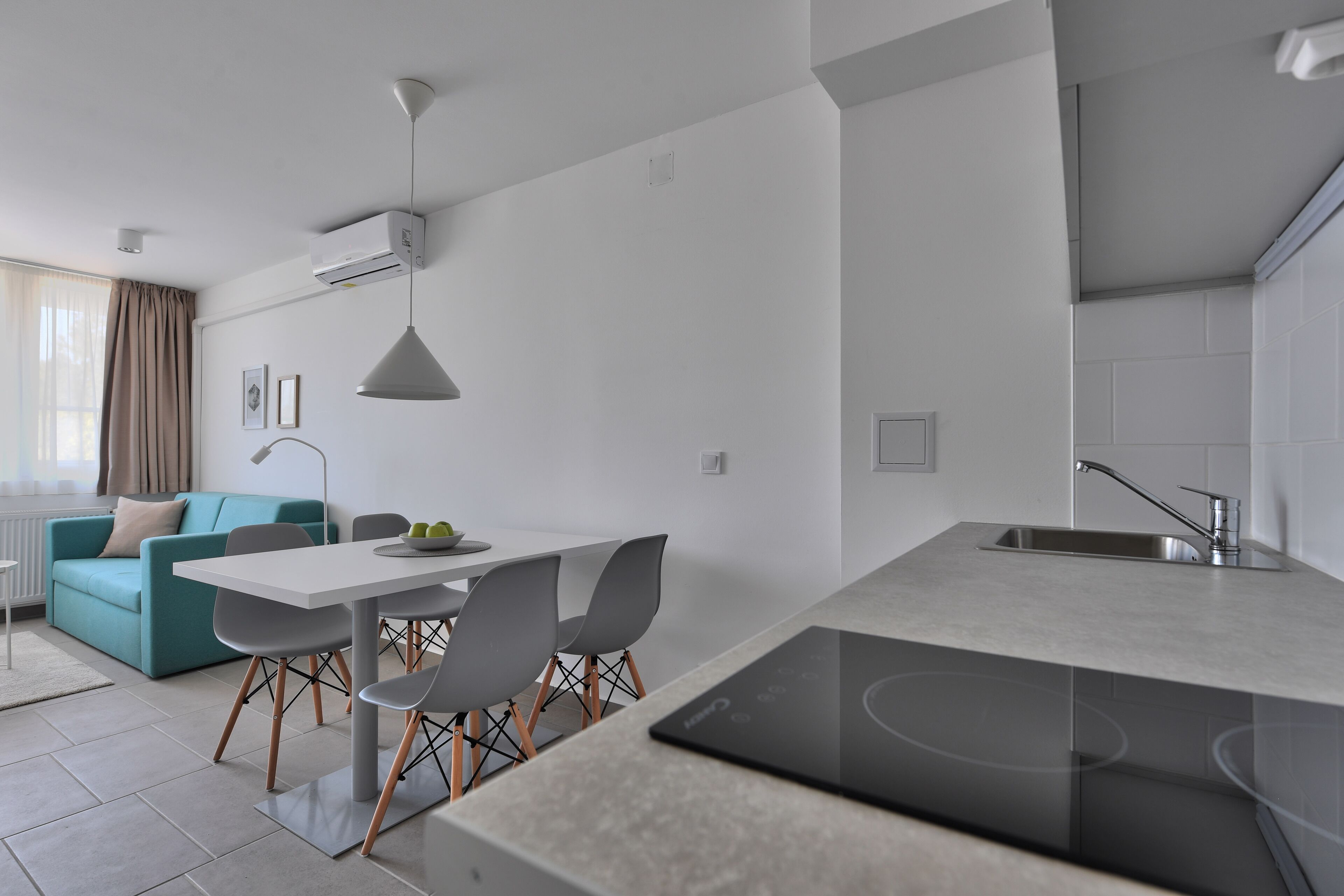 A modern kitchen with a built-in stove, a dining area with a white table and grey chairs, and a teal sofa in the living area.
