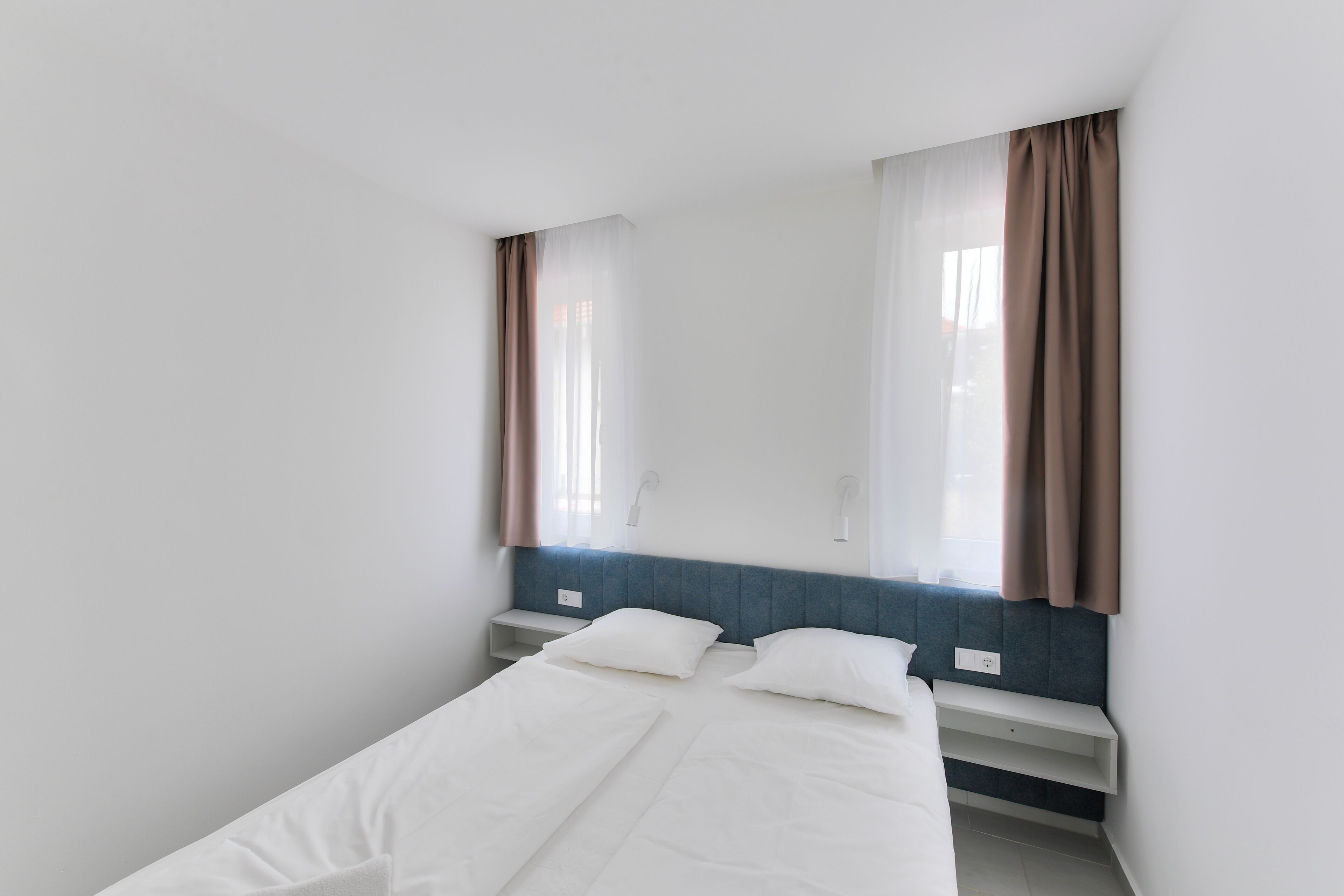A small, single-bed room with a headboard, a window with curtains, and a bedside table.