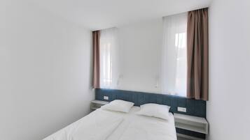 A small, single-bed room with a headboard, a window with curtains, and a bedside table.