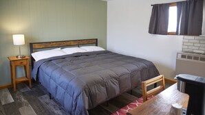 Un dormitorio con cama, una mesita de noche con lámpara, una silla de madera, una mesa con cafetera y una ventana con cortinas.