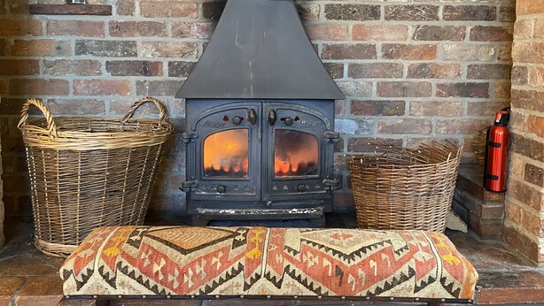 A brick fireplace with a wood-burning stove, two wicker baskets, and a bench with a patterned cushion.