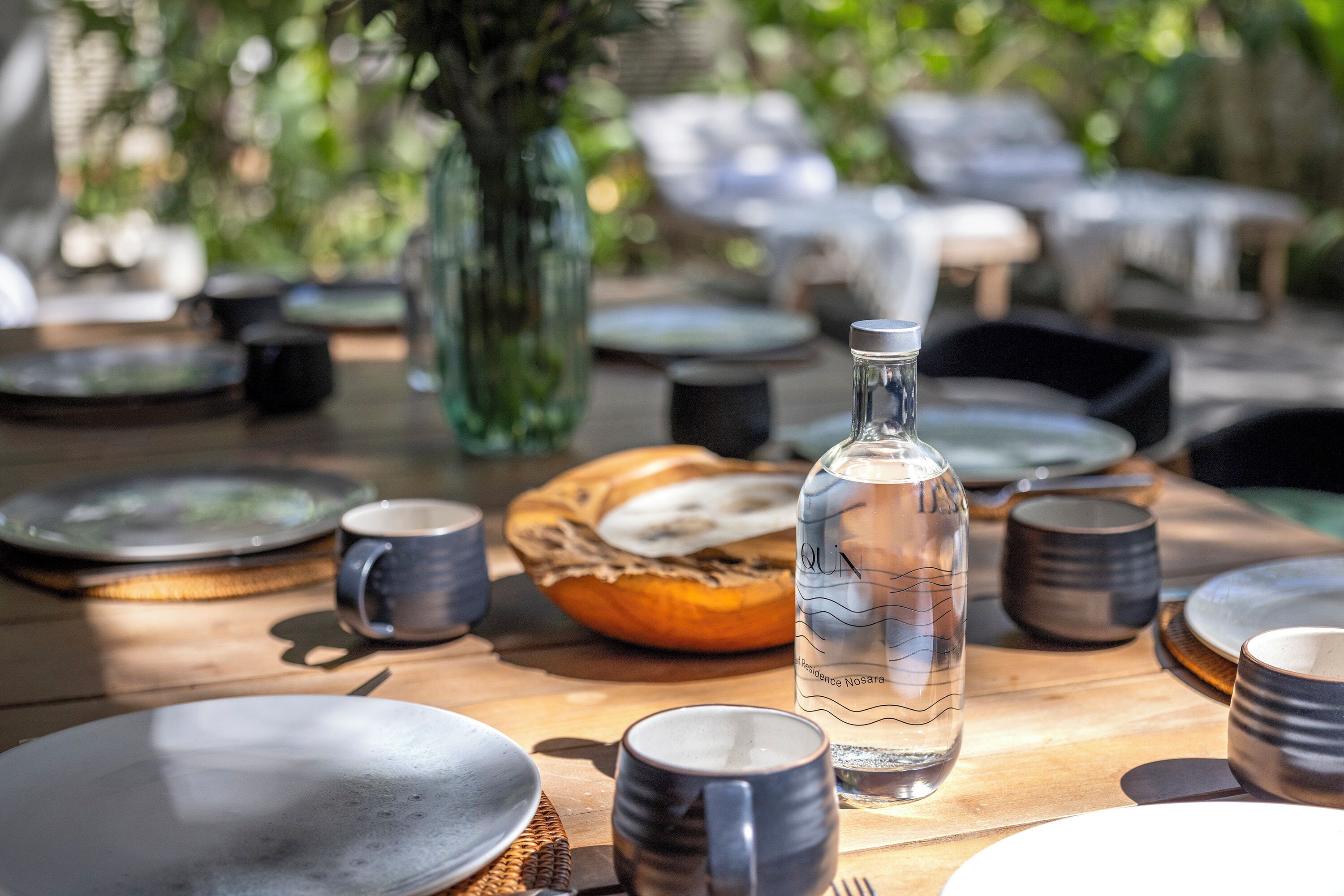 Een eettafel gedekt met borden, mokken en een fles, omringd door stoelen en een mand.