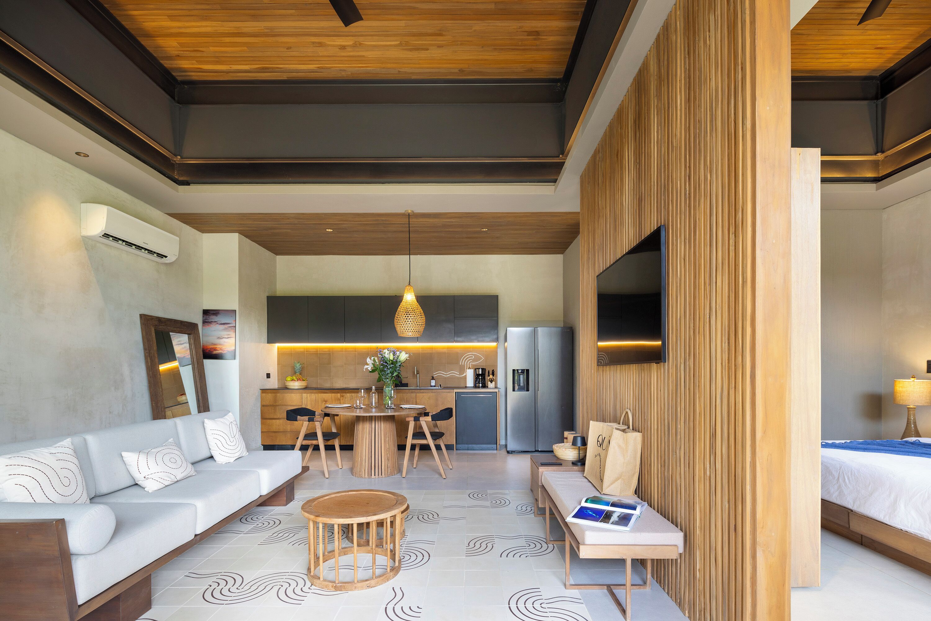 A modern hotel room with a sofa, a dining table, a TV mounted on the wall, and a kitchen area in the background.