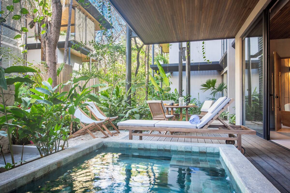 A pool area with lounge chairs and a table, surrounded by greenery and a building with large windows.