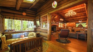 A cozy log cabin interior with a wooden bench, sofa, and armchair, large windows, and a chandelier. - Beautiful open-concept log home in quiet setting on Thompson Lake. (Otisfield)
