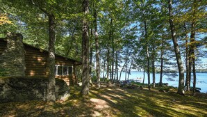 A rustic cabin with a stone foundation, surrounded by tall trees and a lake view. - Beautiful open-concept log home in quiet setting on Thompson Lake. (Otisfield)
