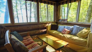 A cozy cabin interior with a wooden sofa, a wooden table, and large windows offering a view of the forest. - Beautiful open-concept log home in quiet setting on Thompson Lake. (Otisfield)