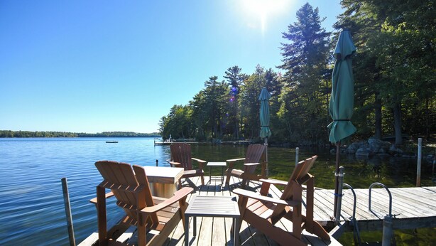 A wooden deck with Adirondack chairs and a table overlooking a lake, surrounded by trees and a clear sky. - Beautiful open-concept log home in quiet setting on Thompson Lake. (Otisfield)