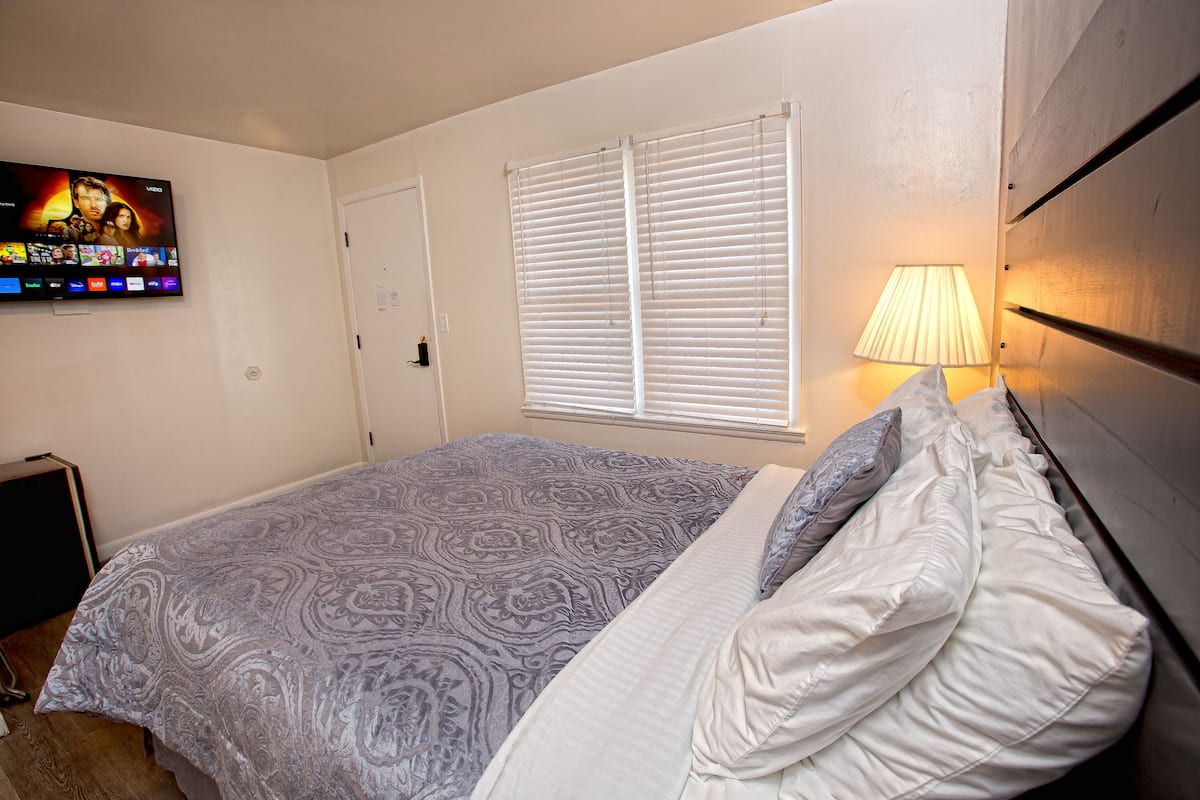 A bedroom with a bed, a TV on the wall, a window with blinds, and a lamp.