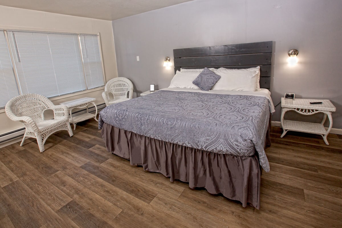 A bedroom with a large bed, two white wicker chairs, a nightstand with a phone, and a window with blinds.