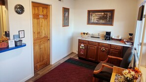 A hotel lobby with a wooden door, a clock, a framed picture, a coffee maker, and a dining area with a chair and table. - Prairie Voyager  (Prairie Du Chien)