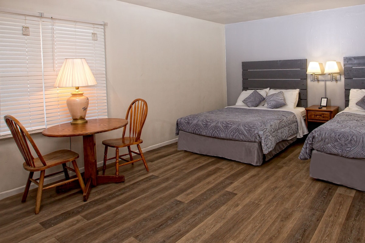 A hotel room with two beds, a wooden table with chairs, a lamp, and a nightstand with two lamps.