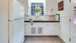 Una cocina con armarios blancos, una ventana con cortinas de cuadros, un lavamanos y un letrero decorativo en la pared.