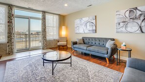 A spacious living room with a large window, a ceiling fan, a grey sofa, a round coffee table, and abstract wall art. - Downtown St Joseph Condo w/ Balcony & Harbor View (St Joseph)