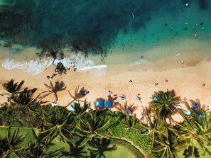 An aerial view of a beach with clear turquoise waters, palm trees, and people sunbathing.