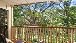 Un balcon doté de balustrades en bois, deux fauteuils confortables, une petite table ronde agrémentée d’une plante, et une vue sur un grand arbre et une végétation luxuriante.