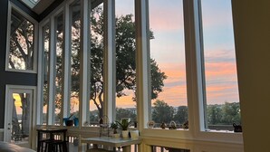 Ein Zimmer mit großen Fenstern, einem Tisch mit Stühlen und Blick auf Bäume und einen Sonnenuntergang.